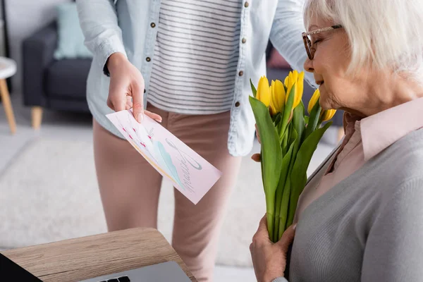 Mujer mayor sosteniendo tulipanes cerca de la hija con tarjeta de felicitación en casa - foto de stock