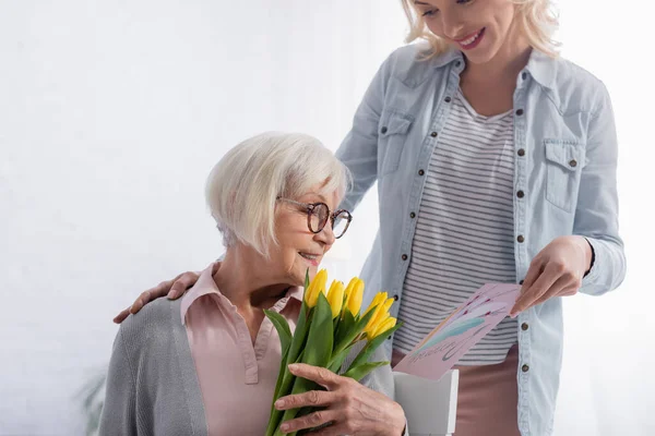 Mujer sonriente sosteniendo tarjeta de felicitación cerca de la madre anciana con tulipanes amarillos - foto de stock