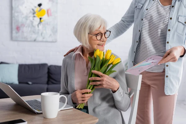Alegre anciana sosteniendo tulipanes cerca de la hija con tarjeta de felicitación y portátil - foto de stock