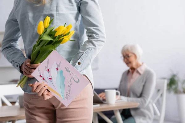 Женщина, прячущая цветы и поздравительную открытку с 8-ю мартовскими буквами рядом с матерью на размытом фоне — стоковое фото