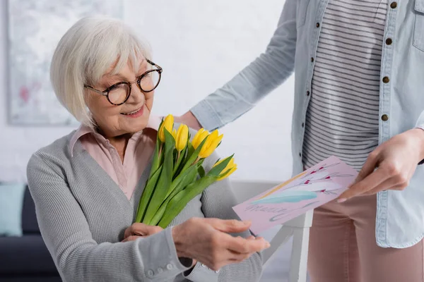 Fröhliche grauhaarige Frau hält Blumen neben Tochter mit Grußkarte zu Hause — Stockfoto