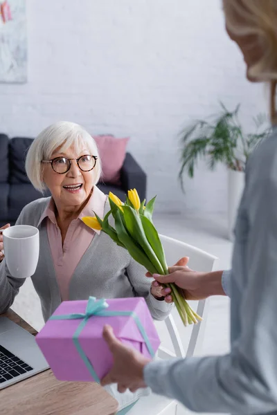 Lächelnde ältere Frau hält Tasse neben Tochter mit Blumen und Geschenk — Stockfoto