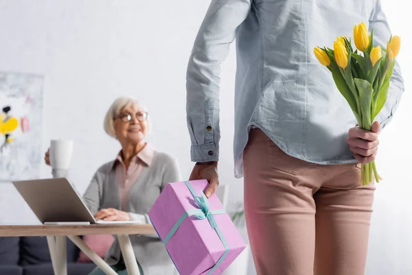 Mujer sosteniendo tulipanes y presente cerca de la madre anciana en sala de estar - foto de stock