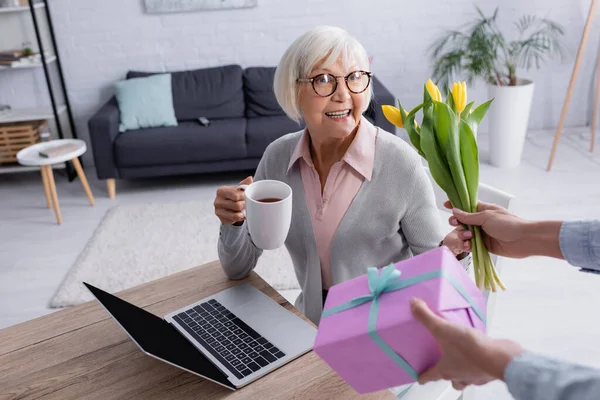 Woman giving flowers and gift box near cheerful mother with tea near laptop — Stock Photo