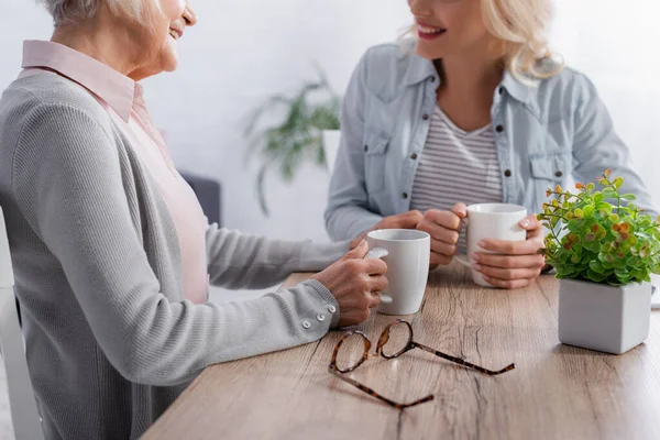 Cropped view of eyeglasses near senior woman holding cup while talking with daughter — Stock Photo