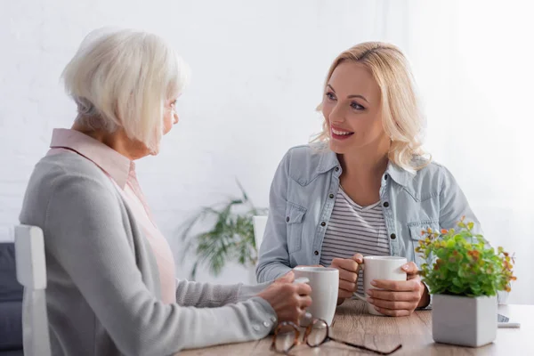 Donna sorridente che tiene la tazza vicino alla madre dai capelli grigi — Foto stock