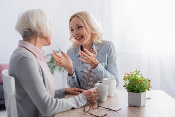 Lächelnde Frau zeigt auf grauhaarige Mutter mit Tasse neben Pflanze und Brille — Stockfoto