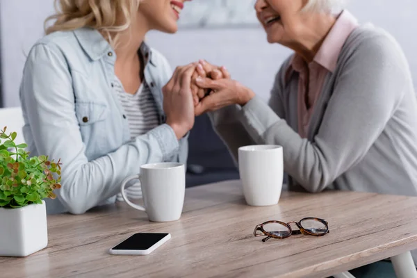 Ausgeschnittene Ansicht von Smartphone, Brille und Tassen neben lächelnder Frau und Mutter auf verschwommenem Hintergrund — Stockfoto