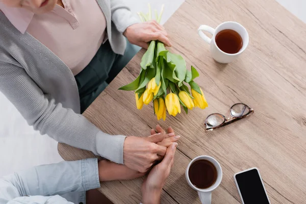 Vue du dessus de la femme âgée avec des fleurs tenant la main de sa fille près du thé et du smartphone — Photo de stock