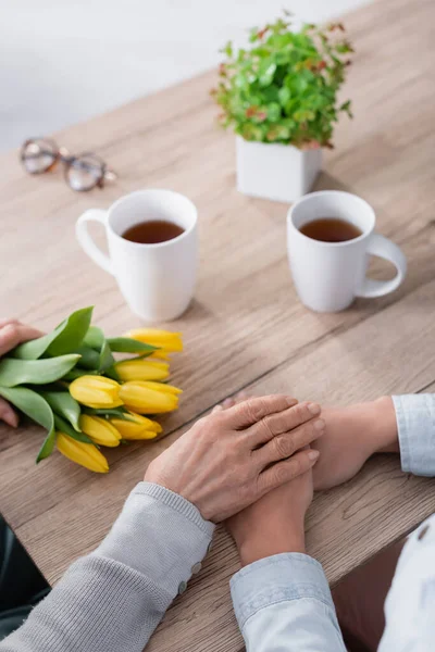 Cropped view of senior woman holding hand of daughter near tulips and cups — Stock Photo