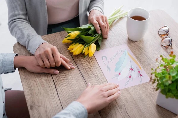 Vista recortada de la mujer mayor sosteniendo la mano de la hija y las flores cerca del té y la tarjeta de felicitación con 8 letras de marzo - foto de stock