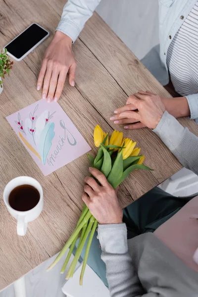 Visão superior da mulher sentada perto do smartphone, cartão de saudação e mãe sênior com tulipas — Fotografia de Stock