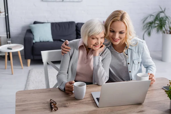 Lächelnde Frau umarmt Mutter mit Tasse neben Laptop auf Tisch — Stockfoto