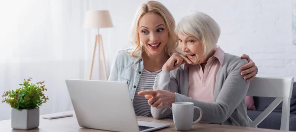 Mujer alegre señalando con el dedo a la computadora portátil cerca del té y la hija, bandera - foto de stock