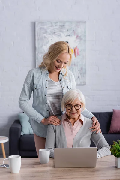 Mujer alegre abrazando a la madre mayor en gafas usando el portátil cerca de copas - foto de stock
