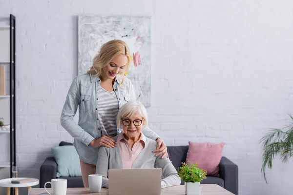 Smiling woman standing near senior mother using laptop in living room — Stock Photo