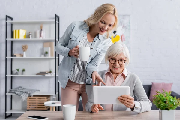 Mujer con la taza apuntando a la tableta digital en la mano de la madre mayor - foto de stock