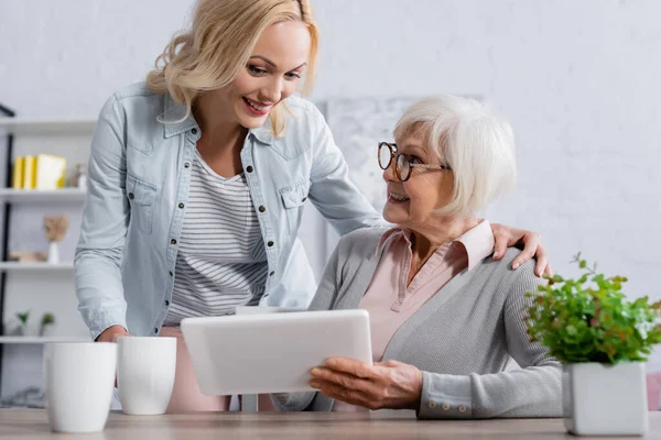 Smiling woman hugging mother with digital tablet near cups on table — Stock Photo