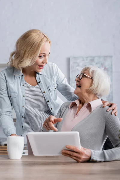 Aufgeregte Frau zeigt auf digitales Tablet in der Nähe von Tochter und Tasse — Stockfoto