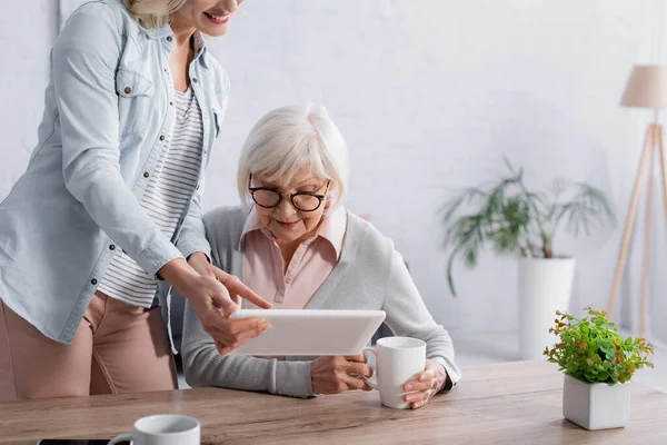 Положительная женщина указывает на цифровой планшет возле пожилой мамы с чашкой дома — стоковое фото