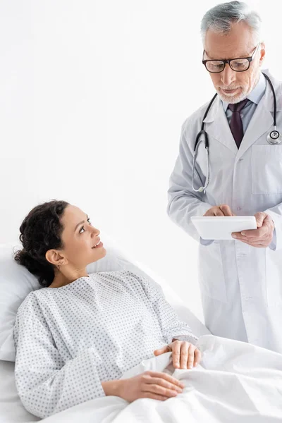 Médico maduro segurando tablet digital perto de mulher afro-americana positiva no hospital — Fotografia de Stock