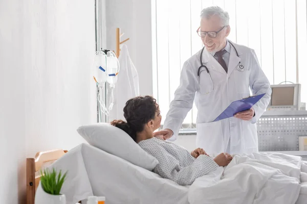 Gioioso medico tenendo appunti mentre tocca spalla di donna afro-americana — Foto stock