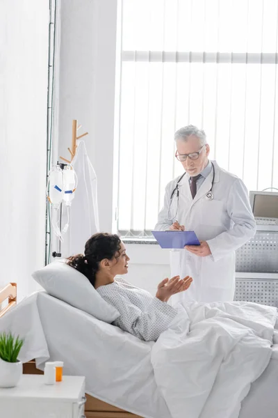 Mujer afroamericana haciendo gestos mientras habla con el médico escribiendo diagnóstico en el portapapeles - foto de stock