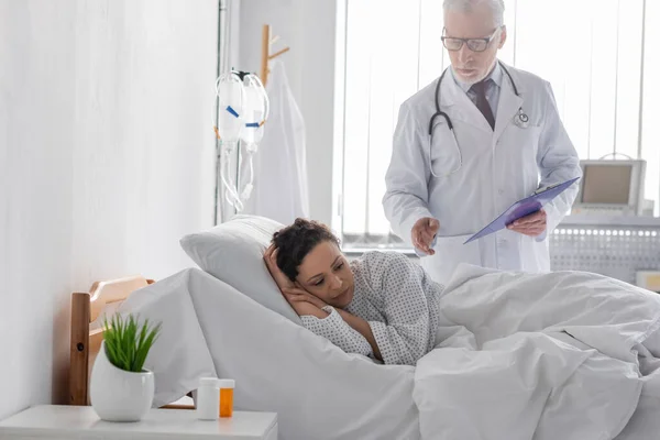 Médico de mediana edad con portapapeles de pie cerca molesto mujer afroamericana acostada en la cama de hospital - foto de stock