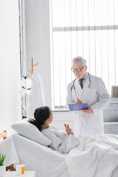 Sonriente doctor escribiendo en portapapeles cerca de afroamericana mujer mostrando pulgares hacia arriba - foto de stock