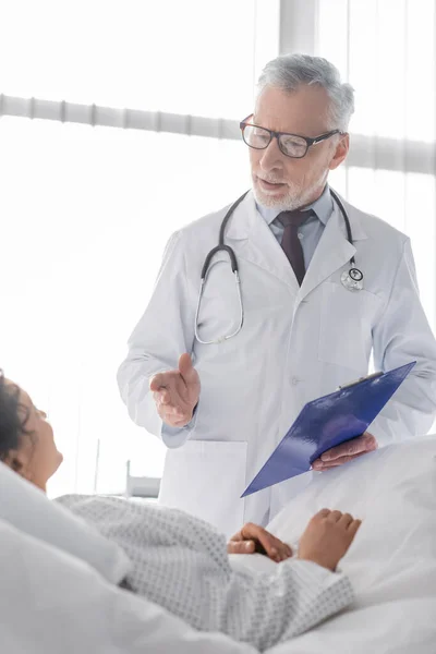 Médico maduro apontando com a mão enquanto conversa com a mulher americana africana em primeiro plano borrado — Fotografia de Stock