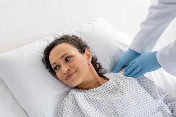 Medico in lattice guanti toccare spalla di sorridente donna africana americana sdraiato nel letto d'ospedale — Foto stock