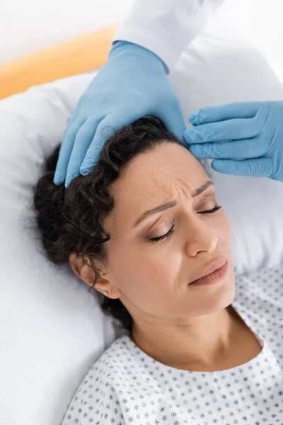 Médico en guantes de látex tocando la cabeza de mujer afroamericana acostada en la cama con los ojos cerrados - foto de stock