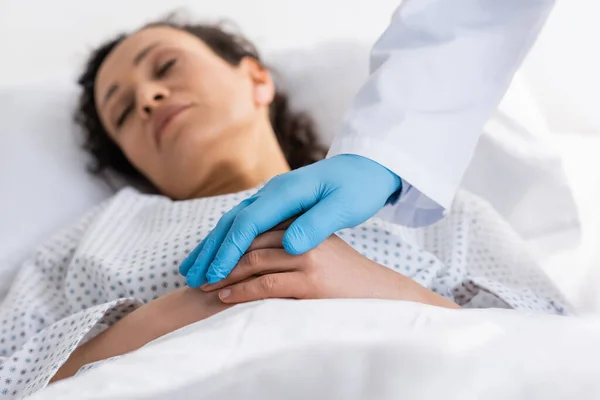 Doctor en guante de látex tocando las manos de la mujer afroamericana dormida en la clínica, fondo borroso - foto de stock