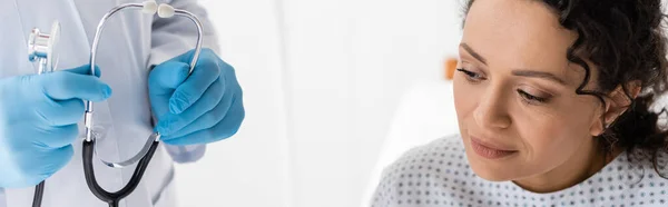 Doctor in latex gloves holding stethoscope near african american woman in clinic, banner — Stock Photo