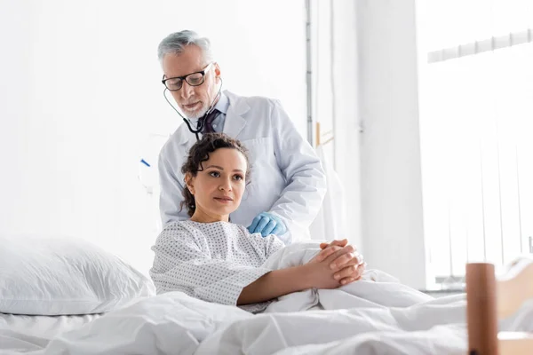 Médico de mediana edad en gafas que examinan mujer afroamericana con estetoscopio en la clínica - foto de stock