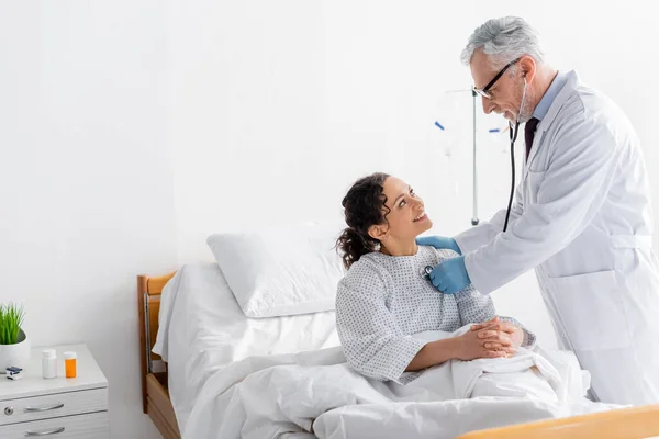 Médico examinando alegre afroamericana mujer con estetoscopio en el hospital - foto de stock