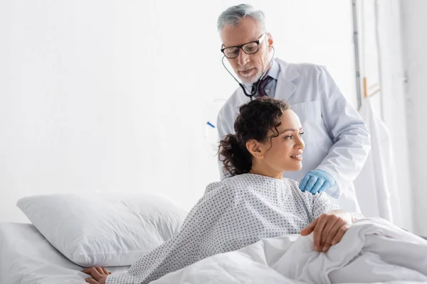 Médico maduro en gafas que examinan mujer afroamericana con estetoscopio en el hospital — Stock Photo