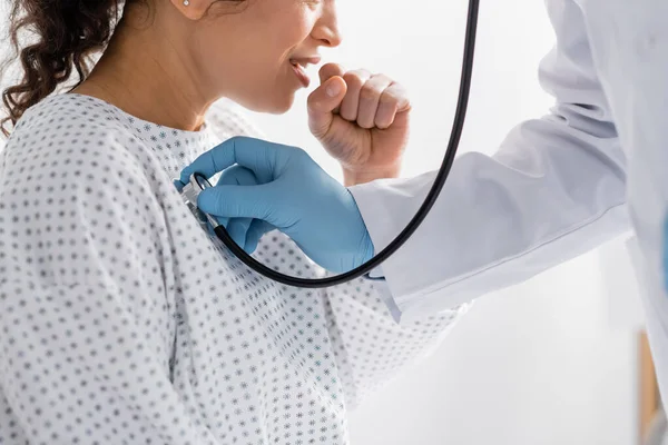 Vista parcial del médico en guantes de látex examinando la tos mujer afroamericana con estetoscopio - foto de stock