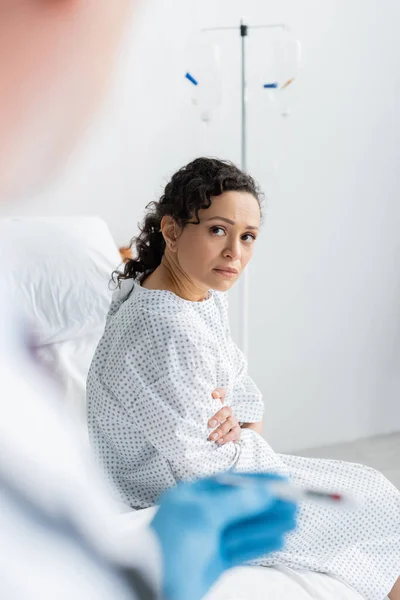 Upset african american woman sitting on hospital bed near doctor with thermometer on blurred foreground — Stock Photo