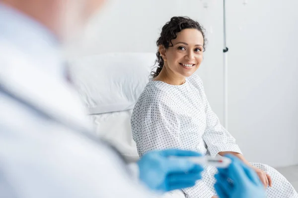 Mulher afro-americana sorrindo perto de médico em luvas de látex segurando termômetro em primeiro plano borrado — Fotografia de Stock