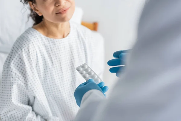 Vista cortada de mulher afro-americana perto de médico em luvas de látex segurando pílulas em primeiro plano borrado — Fotografia de Stock