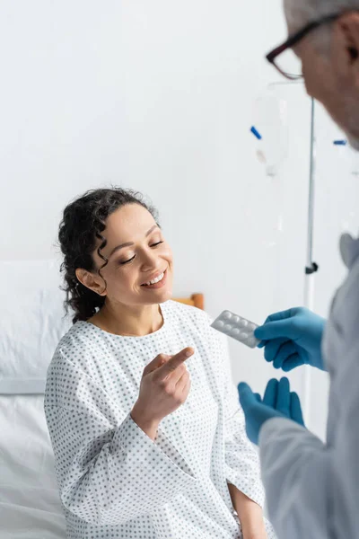 Lächelnde Afroamerikanerin zeigt auf Tabletten in den Händen des Arztes in Latexhandschuhen auf verschwommenem Vordergrund — Stockfoto