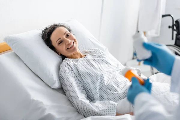 Mujer afroamericana sonriente acostada en la cama de hospital cerca del médico con medicamentos en primer plano borroso - foto de stock