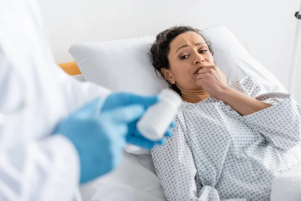 Mujer afroamericana preocupada acostada en la cama de hospital cerca del doctor borroso con guantes de látex apuntando al contenedor con medicamentos - foto de stock