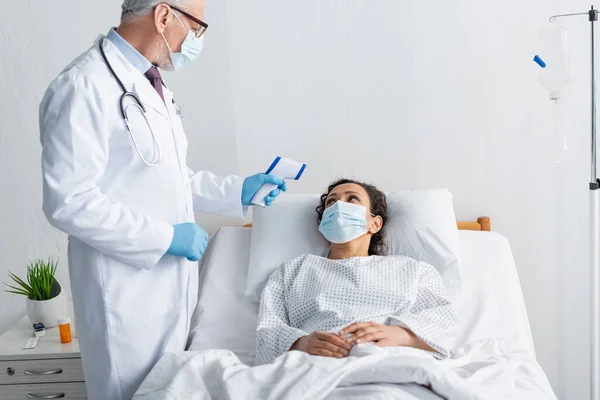 Mature doctor in medical mask measuring temperature of sick african american woman — Stock Photo