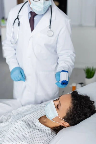 Médecin mesurant la température de la femme afro-américaine en masque médical avec pyromètre — Photo de stock