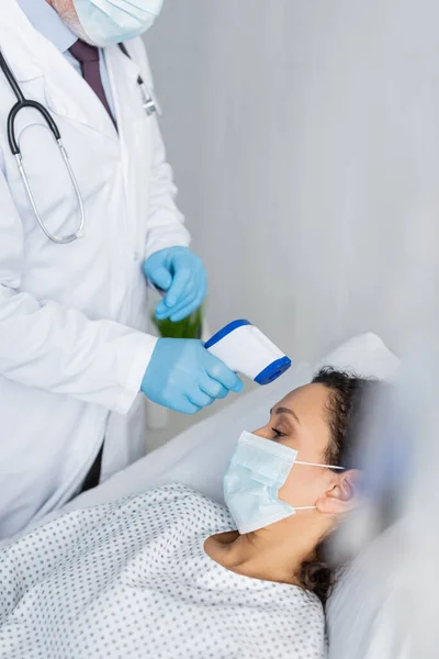 Mujer afroamericana en máscara médica cerca del médico midiendo su temperatura con pirómetro - foto de stock