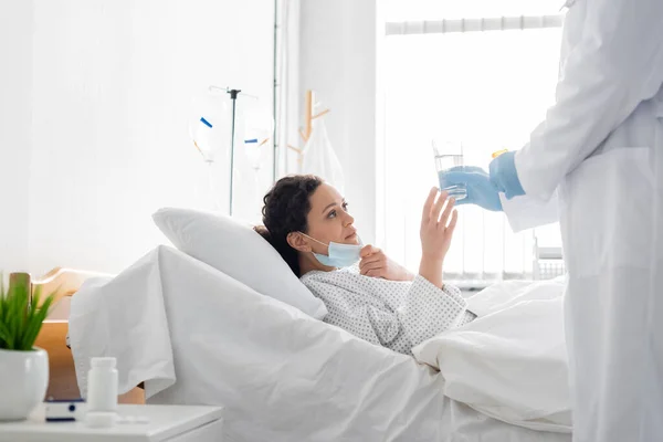 Médico em luvas de látex dando vidro de água para mulher afro-americana doente em cama de hospital — Fotografia de Stock