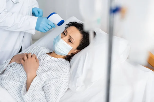 Doctor in latex gloves measuring temperature of african american woman lying in medical mask — Stock Photo
