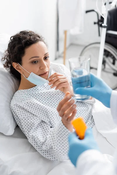 Donna afro-americana che toglie la maschera medica vicino al medico sfocato con farmaci e bicchiere d'acqua — Foto stock